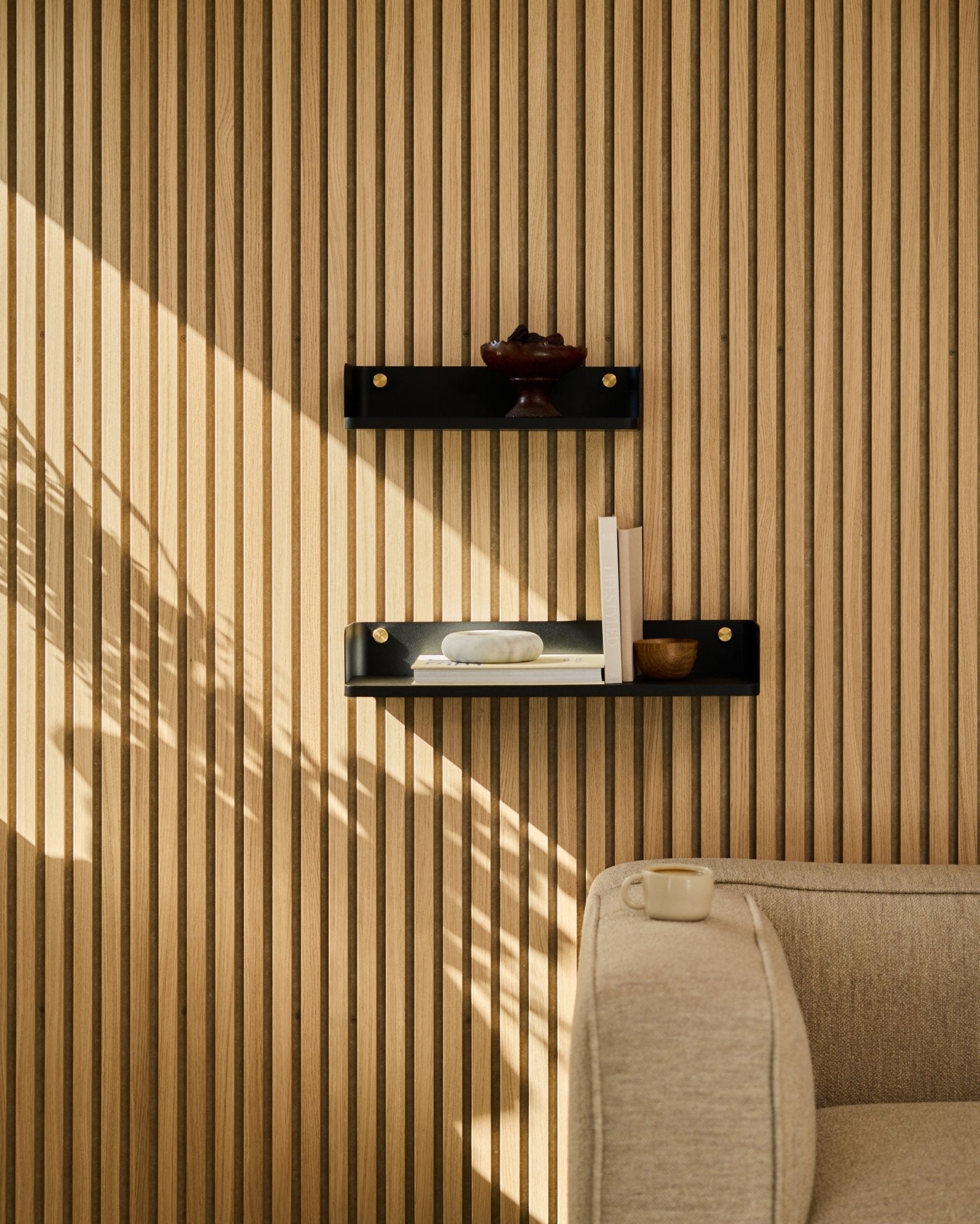Sunlight illuminates a beige sofa and WoodUpps Linn bookshelf in black oak, 55.9 cm, set against a minimalist Nordic slatwall acoustic panel.